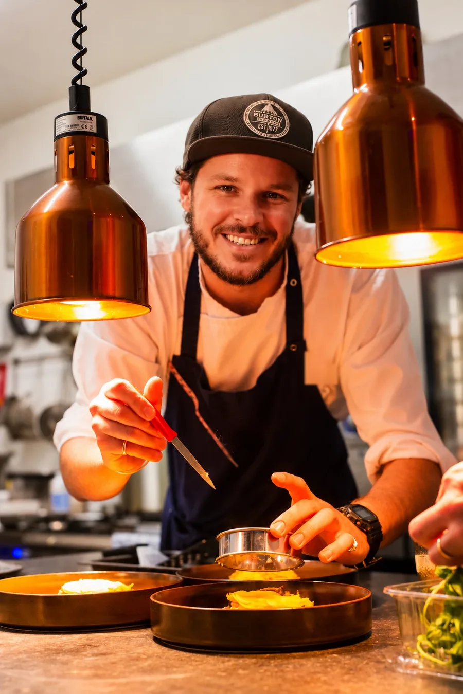 Chef sorrindo enquanto monta um prato na cozinha, sob luminárias de cobre